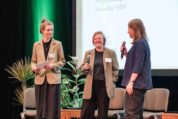 Auf der Bühne von links: Moderatorin Louisa Schmidt sowie die Baukultur NRW-Projektmanagerinnen Fenna Tinnefeld und Annika Stremmer, die den Kongress Grün! Blau! Grau! im Saalbau Witten organisiert haben. Foto: Sebastian Becker